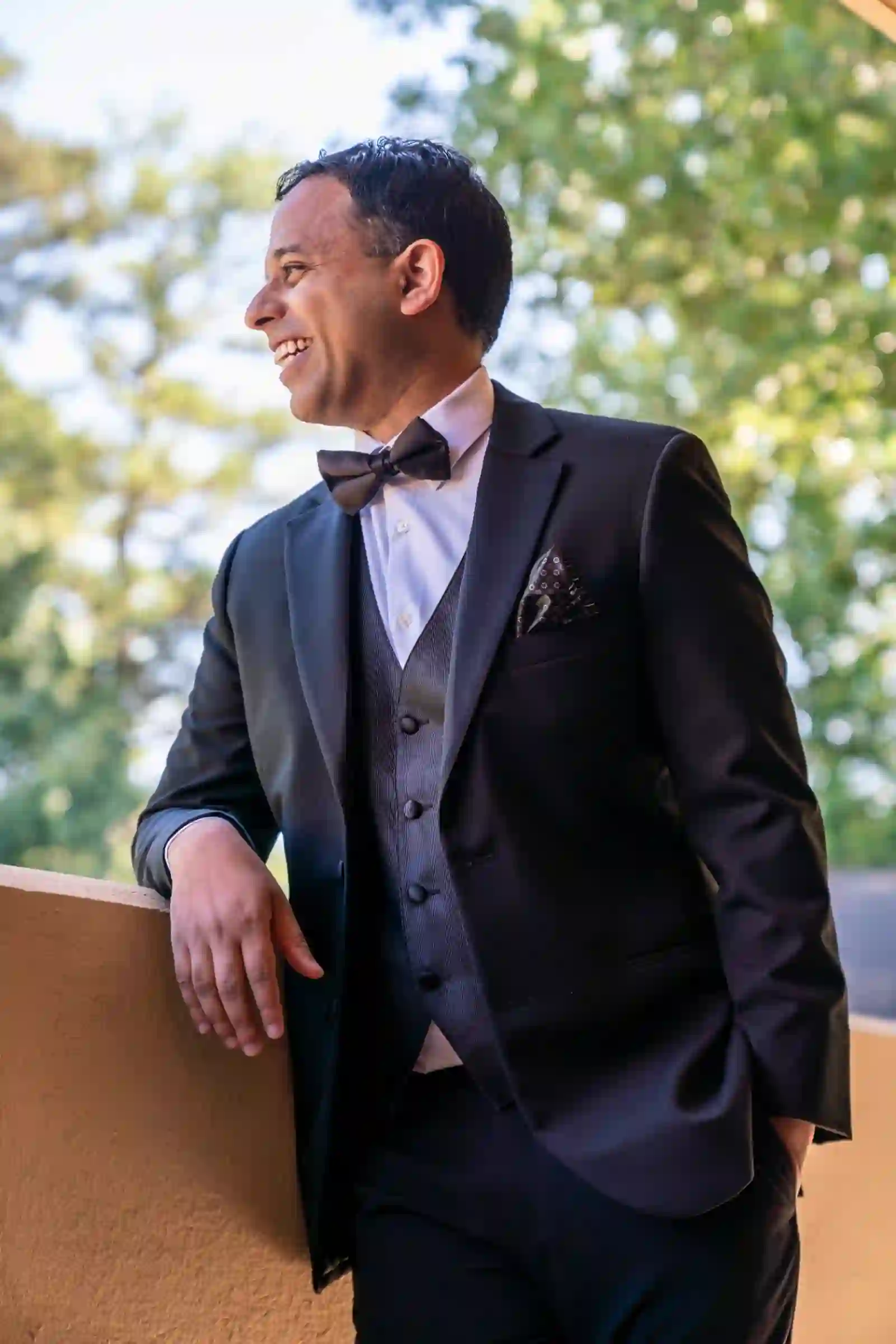 Groom in a tuxedo and bow tie laughing on a sunlit balcony