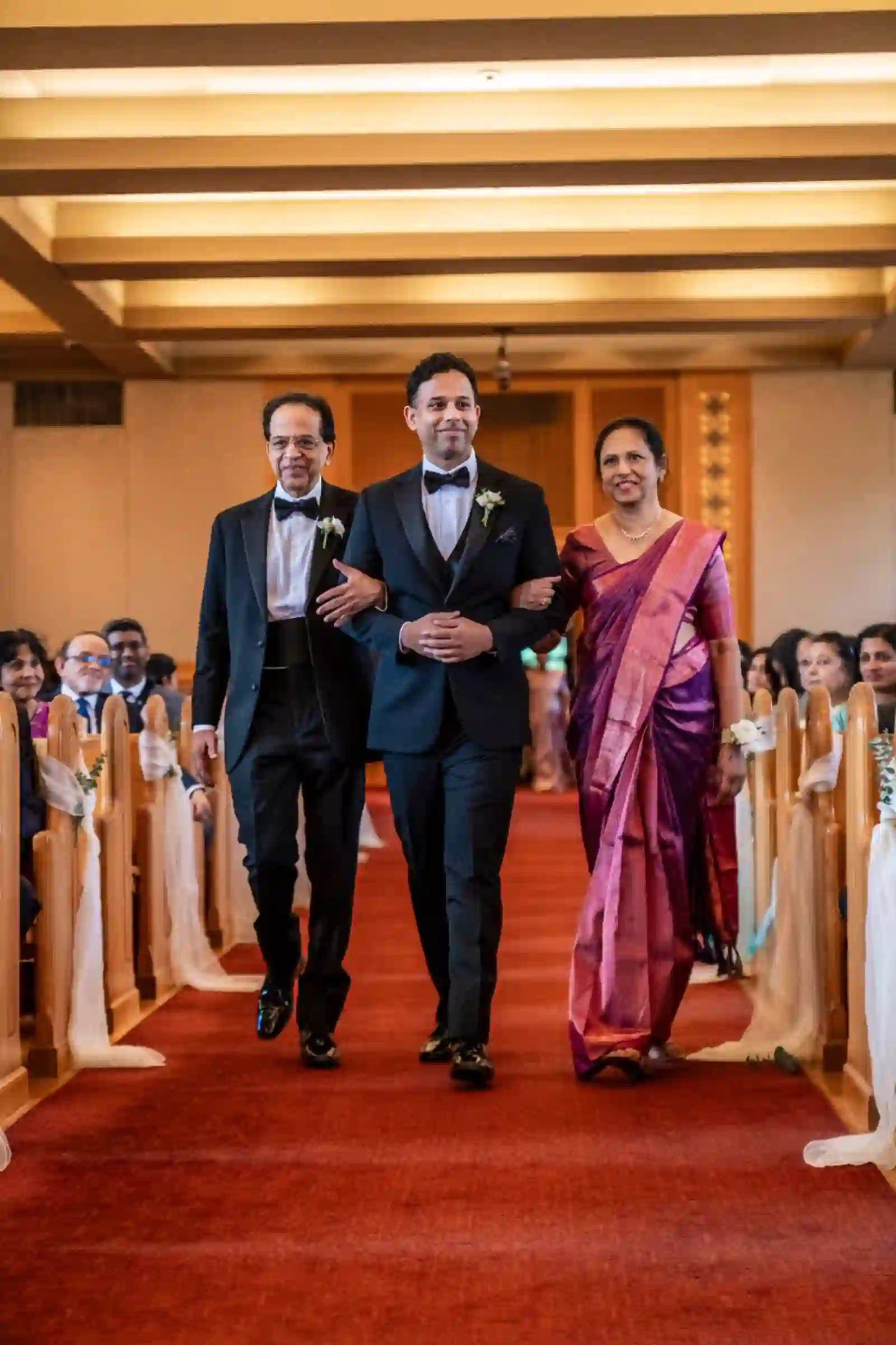 Groom walking down the aisle between his parents, his mother in a magenta sari