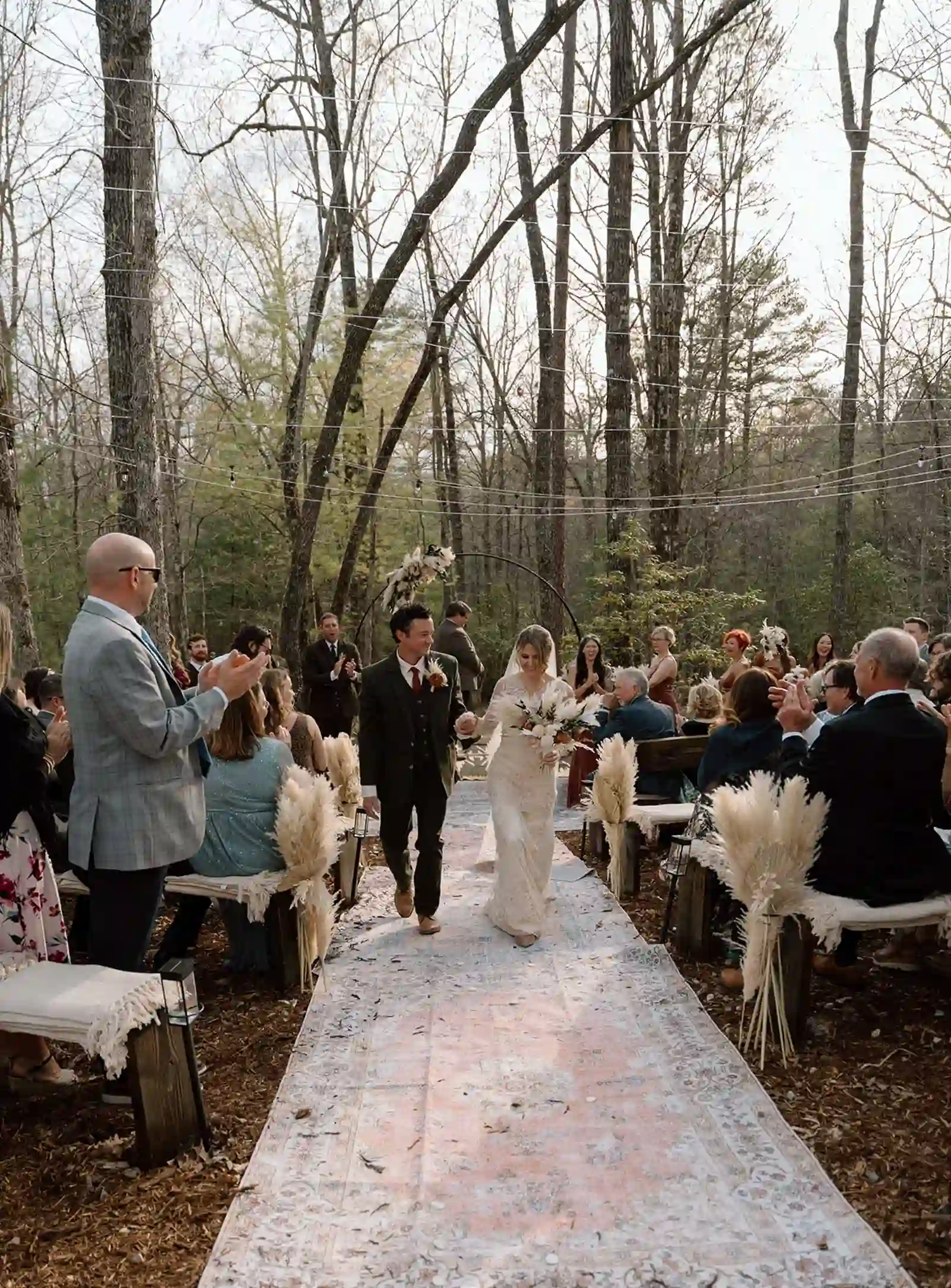 Bride and groom recessing down a vintage rug aisle as guests applaud in the pines