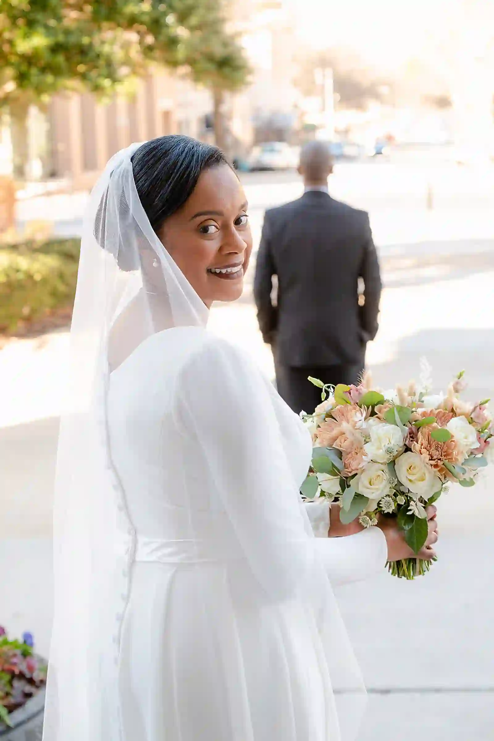Bride glancing over her shoulder with a soft veil and pastel bouquet on a sunlit downtown sidewalk