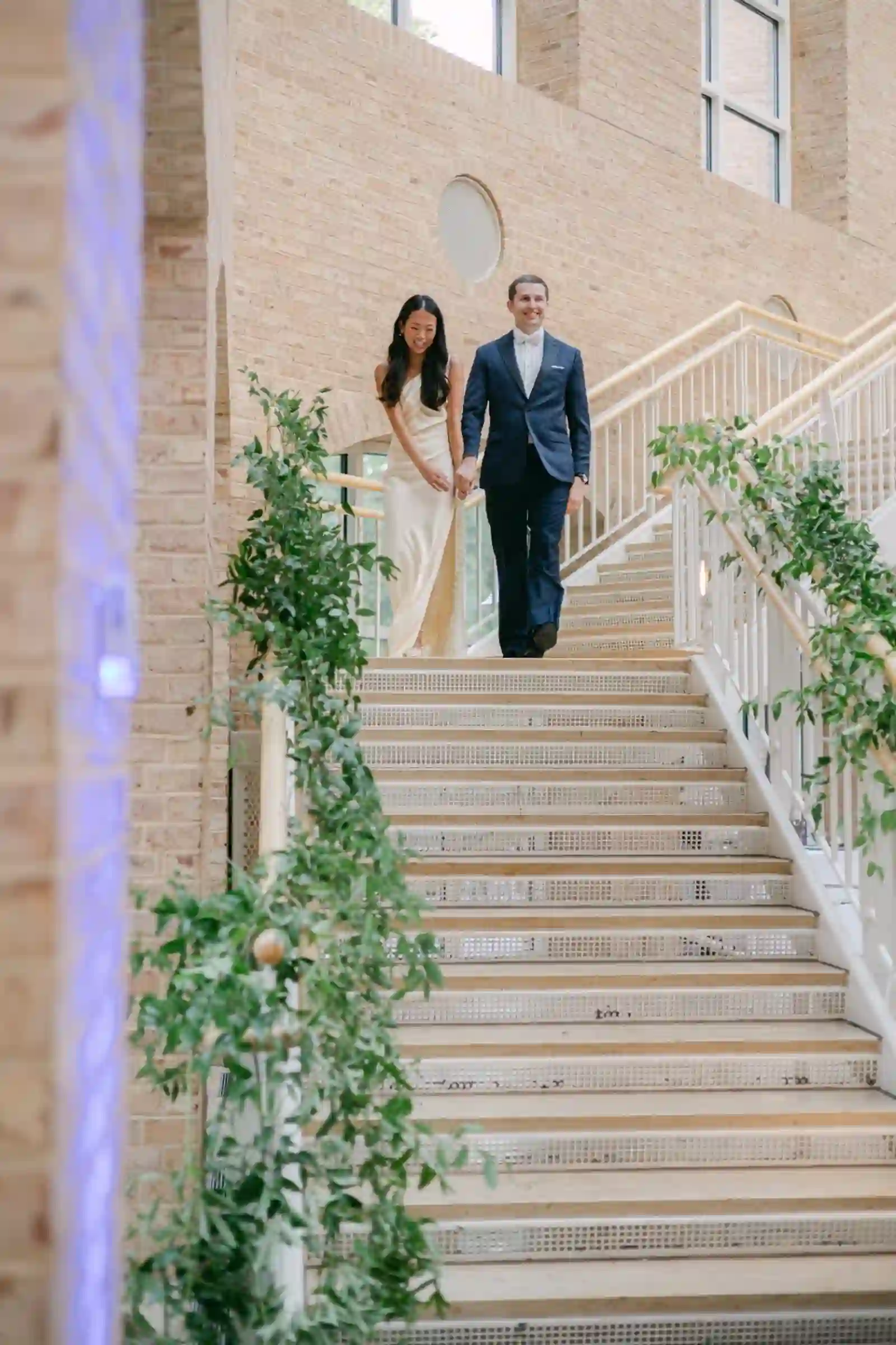 Bride in a silk gown and groom in navy walking down a greenery-draped grand staircase