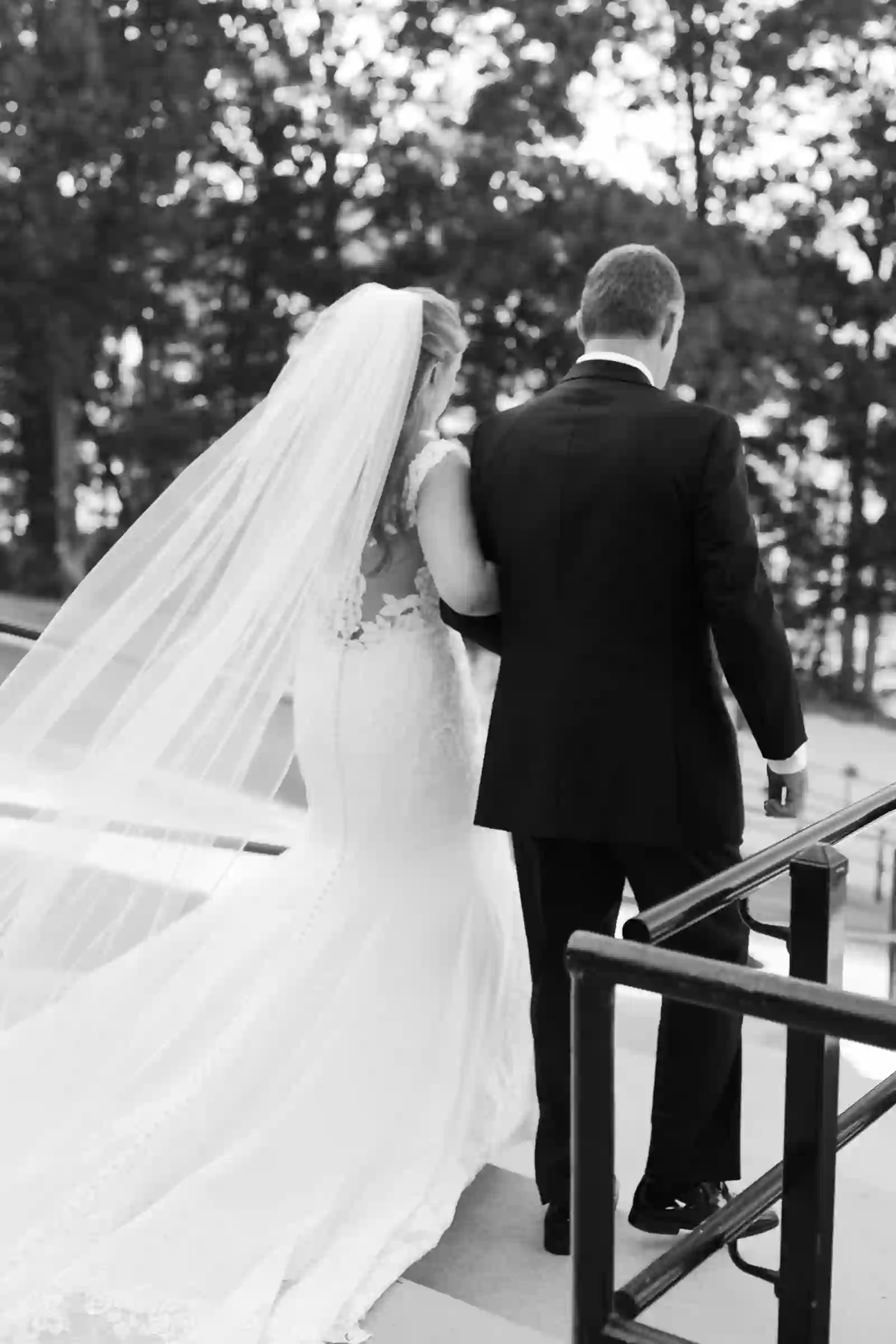 Black and white of bride and father walking arm in arm, veil trailing behind