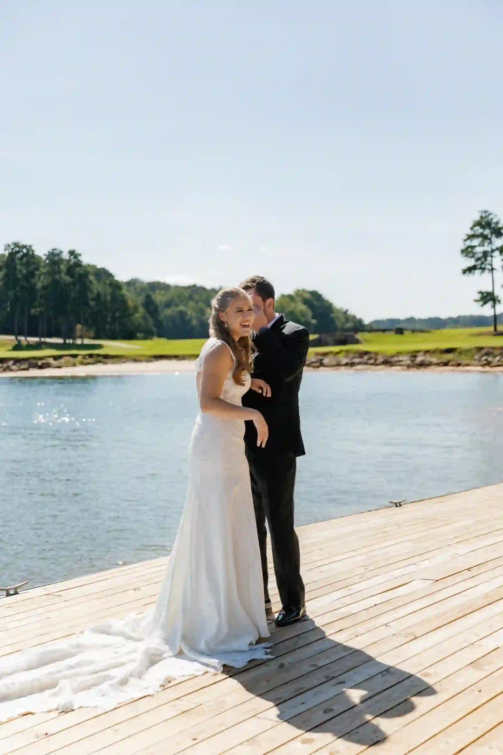 Bride laughing as her groom whispers into her ear by the water