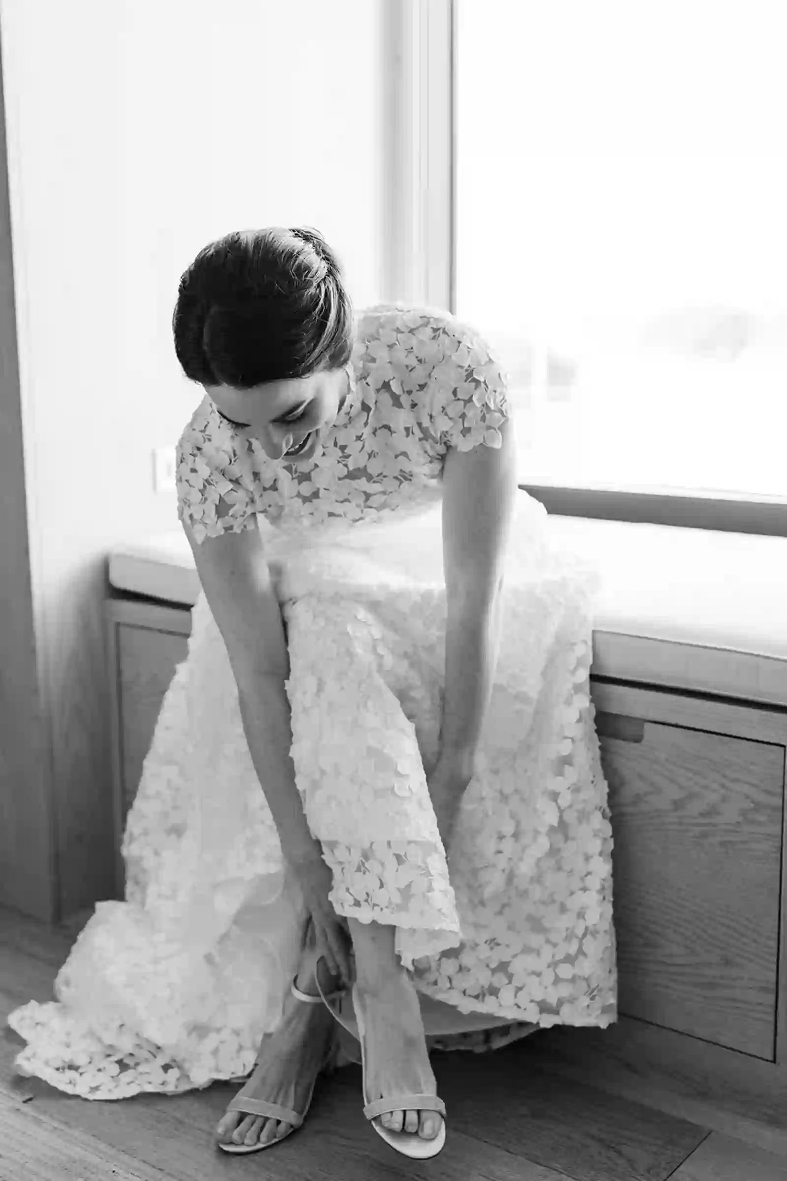 Black and white portrait of the bride adjusting her shoes by the window