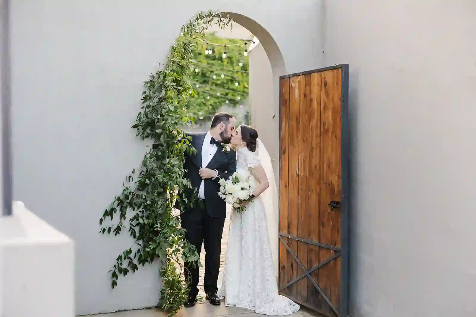 Bride and groom kissing under a stucco arch with a wooden door and string lights